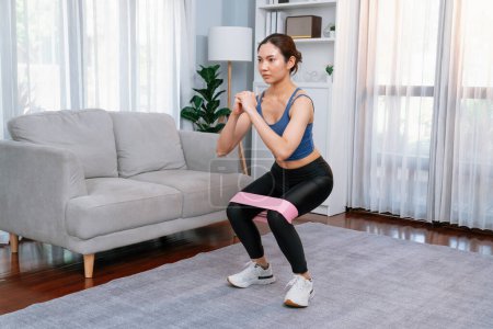 Photo for Vigorous energetic woman doing exercise at home with resistance sport band for leg muscle gain. Young athletic asian woman strength and endurance training session as home workout routine with squat. - Royalty Free Image