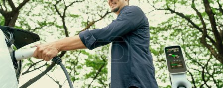 Photo for Man recharging battery for electric car during road trip travel EV car in natural forest or national park. Eco friendly travel during vacation and holiday. Panorama banner Exalt - Royalty Free Image