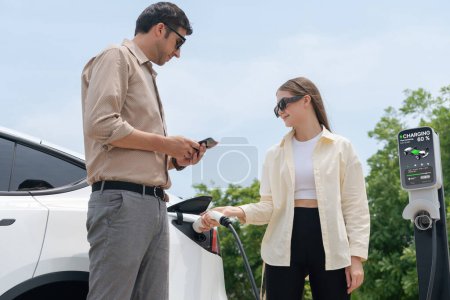 Photo for Young couple use smartphone to pay for electricity at public EV car charging station green city park. Modern environmental and sustainable urban lifestyle with EV vehicle. Expedient - Royalty Free Image