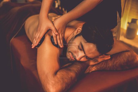 Téléchargez les photos : Massage aux pierres chaudes au salon de spa dans un complexe de luxe avec lumière chaude aux bougies, client homme heureux profitant d'un massage aux pierres de basalte spa glisse sur le corps avec une chaleur apaisante. Tranquillité - en image libre de droit