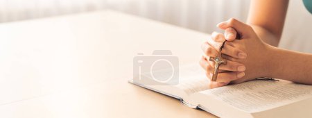 Téléchargez les photos : Image recadrée d'une femme lisant un livre de bible tout en tenant la croix à une table en bois avec un fond flou. Concept d'espérance, de religion, de foi, de christianisme et de bénédiction divine. Chaud. Émergence. - en image libre de droit