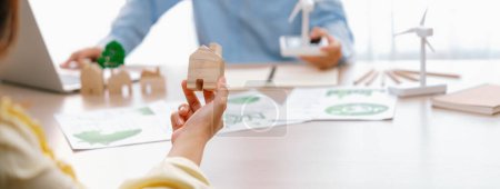 Photo for Cropped image of businesswoman presents eco-friendly house by using green design to manager while holding the wooden house block at table with windmill model placed with document. Delineation. - Royalty Free Image