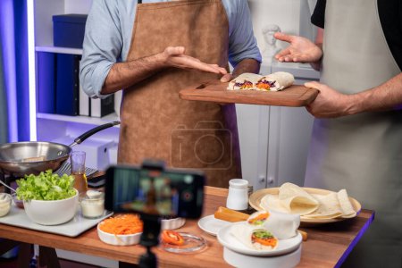 Foto de Vista recortada de los influencers chef presentando rollo de ensalada fresca en la cocina paso a paso, streaming a través de teléfono inteligente en las redes sociales canal en vivo, mostrando ingredientes envoltura de tortilla suave vegetal. Vendible. - Imagen libre de derechos