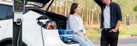Photo for Panorama banner couple recharging battery for electric car while relaxing and sit on the trunk during road trip travel EV car in autumnal forest. Eco friendly travel on vacation during autumn. Exalt - Royalty Free Image