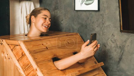 Foto de Un retrato de una hermosa mujer caucásica jugando su teléfono móvil mientras usa un gabinete de sauna de madera en tono cálido. Atractiva hembra con hermosa piel tomando una foto. Fondo gris. Tranquilidad - Imagen libre de derechos
