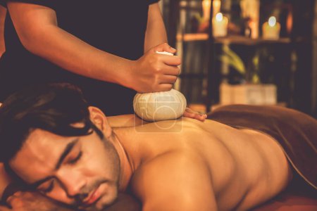 Photo for Hot herbal ball spa massage body treatment, masseur gently compresses herb bag on man body. Tranquil and serenity of aromatherapy recreation in warm lighting of candles at spa salon. Quiescent - Royalty Free Image