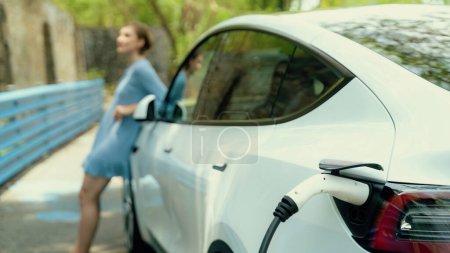 Photo for Focus EV charger by electric charging station recharging car on blurred background of beautiful young woman checking cars battery from smartphone. Eco-friendly travel with EV car. Panorama Perpetual - Royalty Free Image