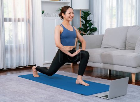 Téléchargez les photos : Femme asiatique en vêtements de sport faisant de l'exercice de yoga sur tapis de fitness comme sa routine d'entraînement à domicile. Mode de vie sain de soins du corps femme regarder en ligne la vidéo de yoga sur ordinateur portable. Vigoureux - en image libre de droit