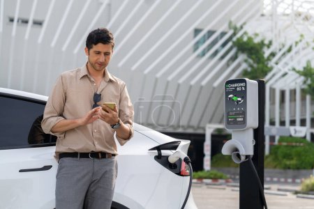 Photo for Young man use smartphone to pay for electricity at public EV car charging station at city commercial mall parking lot. Modern environmental and sustainable urban lifestyle with EV vehicle. Expedient - Royalty Free Image