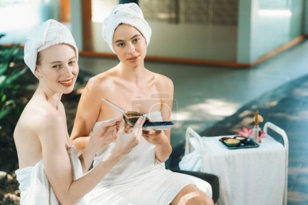 Foto de Un retrato de dos chicas de spa en toalla blanca mirando a la cámara mientras sostiene la máscara facial casera al aire libre rodeada de un entorno natural tranquilo. Concepto saludable y de belleza. Tranquilidad. - Imagen libre de derechos