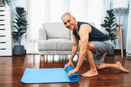 Téléchargez les photos : Homme âgé actif et sportif se préparant, tapis d'exercice de fitness roulant sur le plancher du salon à la maison. L'exercice à domicile comme concept d'un mode de vie sain après la retraite. Clout - en image libre de droit