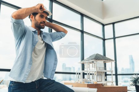 Photo for Portrait of skilled architect engineer wear safety helmet while sitting on table with house model, project plan and architectural equipment surrounded by skyscraper view. Civil engineering. Tracery. - Royalty Free Image