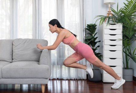 Téléchargez les photos : Fit jeune femme asiatique étirant sa jambe son corps à l'aide d'un canapé dans le salon. Mode de vie sain entraînement routine d'entraînement à la maison. Concept d'exercice d'équilibre et de flexibilité. Vigoureux - en image libre de droit