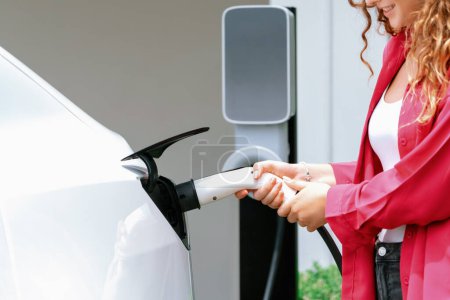 Photo for Modern eco-friendly woman recharging electric vehicle from home EV charging station. EV car technology utilized for home resident to future environmental sustainability. Synchronos - Royalty Free Image