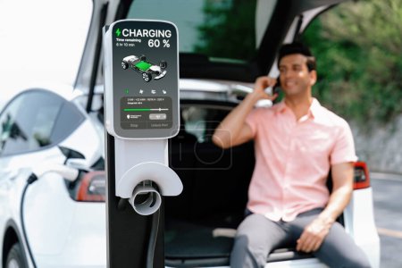 Photo for EV car recharging battery from outdoor EV charger display battery status on blur background of man using smartphone with natural scenic as concept of eco-friendly travel with clean energy. Perpetual - Royalty Free Image
