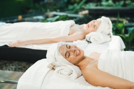 Téléchargez les photos : Deux belles jeunes femmes allongées sur un lit de spa avec serviette blanche tout en abattant dans une profonde détente arrondie par une nature relaxante et apaisante. Concept santé et beauté. Fond flou. Tranquillité. - en image libre de droit
