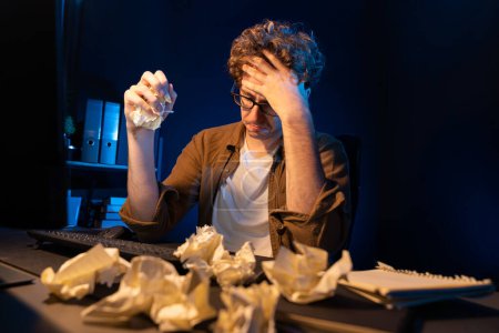 Photo for Stressful creative manager working with overworked project surrounded crumpled papers while waiting email sending back on laptop screen in casual shirt at night time at neon modern office. Gusher. - Royalty Free Image