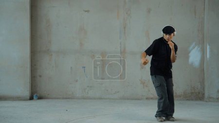 Handsome hipster practicing break dancing while listening music from headphone with cement background. Motion shot of street dancer perform robot dance. Outdoor sport 2024. Modern dance. Endeavor.