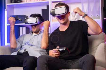 Téléchargez les photos : Copain gamers jouer à des jeux vidéo à l'aide de joysticks et VR casque de la technologie virtuelle en réalité dans la salle de studio avec néon lumière bleue. Vivre confortablement à l'intérieur avec gagnant de combat joyeux. Vendu. - en image libre de droit