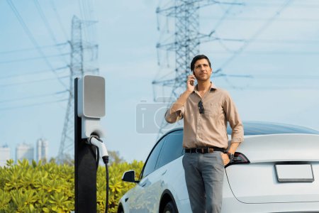 Téléchargez les photos : Homme parlant sur le téléphone tout en rechargeant la batterie de voiture EV à la station de charge connectée à la tour de réseau électrique comme industrie électrique pour une utilisation de voiture respectueuse de l'environnement. - en image libre de droit