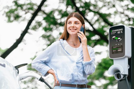 Téléchargez les photos : Jeune femme utilisant smartphone application bancaire en ligne pour payer la charge de la batterie de voiture électrique à partir de la station de charge EV pendant les vacances voyage sur la route dans le parc national ou la forêt d'été. Exalt. - en image libre de droit