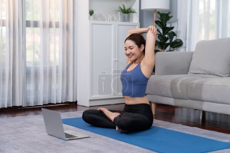 Photo for Asian woman in sportswear doing yoga exercise on fitness mat as her home workout training routine. Healthy body care lifestyle woman watching online yoga video on laptop. Vigorous - Royalty Free Image