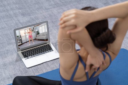 Foto de Mujer asiática en ropa deportiva haciendo ejercicio de yoga en la alfombra de fitness como su rutina de entrenamiento en casa. Cuidado del cuerpo saludable estilo de vida mujer viendo video de yoga en línea en el ordenador portátil. Vigoroso - Imagen libre de derechos