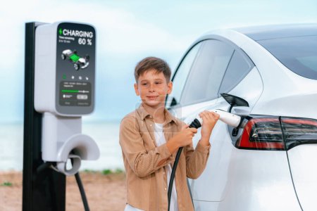 Photo for Little boy recharging eco-friendly electric car from EV charging station. EV car road trip travel by the seashore by alternative vehicle powered by clean renewable and sustainable energy. Perpetual - Royalty Free Image