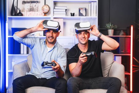 Téléchargez les photos : Copain gamers jouer à des jeux vidéo à l'aide de joysticks et VR casque de la technologie virtuelle en réalité dans la salle de studio avec néon lumière bleue. Vivre confortablement à l'intérieur avec gagnant de combat joyeux. Vendu. - en image libre de droit