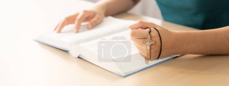 Photo for Cropped image of female reading a bible book while holding cross at wooden table with blurring background. Concept of hope, religion, faith, christianity and god blessing. Warm. Burgeoning. - Royalty Free Image