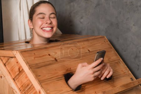 Téléchargez les photos : Un portrait de la magnifique femme caucasienne jouant son téléphone portable tout en utilisant une armoire de sauna en bois dans un ton chaud. Jolie femelle avec une belle peau prenant une photo. Fond gris. Tranquillité - en image libre de droit