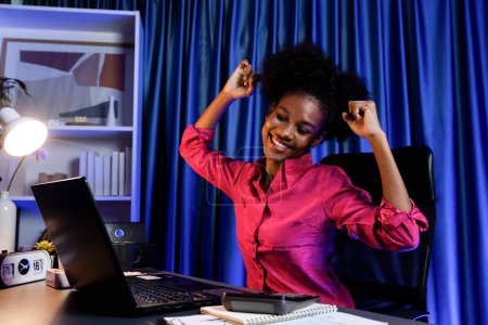 Téléchargez les photos : Femme africaine qui travaille avec un visage heureux rayonnant et souriant, obtenir un nouveau projet d'emploi avec de bonnes affaires ou des informations de bourse de cours de marketing sur écran d'ordinateur portable. Concept d'expression joyeuse. Tastemaker. - en image libre de droit