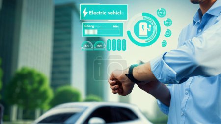Téléchargez les photos : Homme d'affaires vérifier l'état de la batterie de voiture EV sur la recharge hologramme smartwatch de la station de charge ESG parc vert. Style de vie urbain moderne et utilisation écologique de la voiture électrique pour réduire le CO2. Pertinence - en image libre de droit