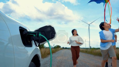 Téléchargez les photos : Focus voiture électrique écologique rechargeant la batterie de la station de recharge à l'aide d'éoliennes renouvelables et propres avec une famille heureuse en cours d'exécution sur la route et jouer avec le cerf-volant en arrière-plan. Pertinence - en image libre de droit