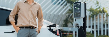 Photo for Young man recharge electric cars battery from charging station in city commercial parking lot. Rechargeable EV car for sustainable environmental friendly urban travel. Panorama Expedient - Royalty Free Image