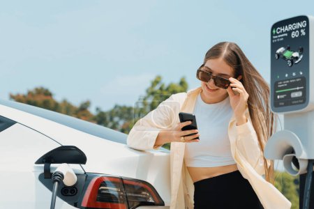 Photo for Young woman use smartphone to pay for electricity at public EV car charging station green city park. Modern environmental and sustainable urban lifestyle with EV vehicle. Expedient - Royalty Free Image