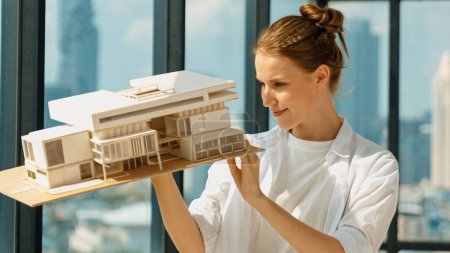 Photo for Young smart caucasian beautiful architect engineer raised house model while inspect carefully surrounded by city skyscraper view. Civil engineering, architectural studio. project planing. Tracery - Royalty Free Image