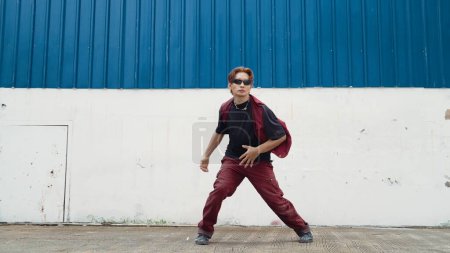 Photo for Handsome street dancer practicing break dancing at white back ground. Sport man wearing hip hop style while moving footsteps to music at street with blue background. Outdoor sport 2024. Endeavor. - Royalty Free Image