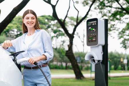 Photo for Young woman recharge EV electric vehicles battery from EV charging station in outdoor green city park scenic. Eco friendly urban transport and commute with eco friendly EV car travel. Exalt - Royalty Free Image
