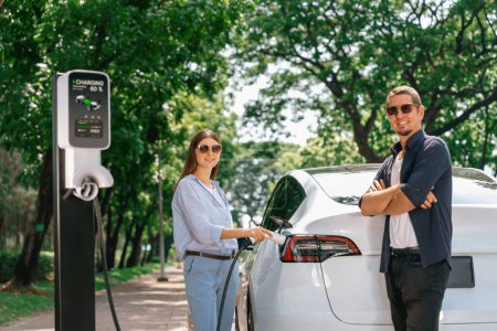 Photo for Lovely young couple wearing sun glasses recharging battery for electric car during road trip travel EV car in natural forest or national park. Eco friendly travel during vacation and holiday. Exalt - Royalty Free Image