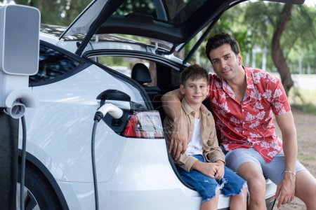 Téléchargez les photos : Vacances de voyage en famille avec véhicule électrique, père et fils rechargent voiture EV avec l'énergie verte et propre. Nature et voyage avec une voiture écologique pour un environnement durable. Perpétuel - en image libre de droit