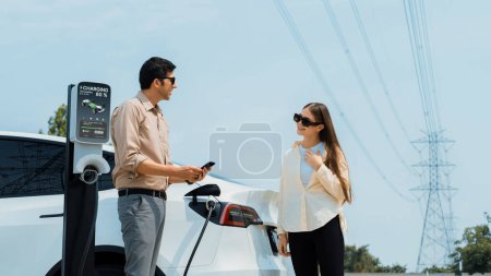 Photo for Couple pay for electricity with smartphone while recharge EV car battery at charging station connected to power grid tower electrical as electrical industry for eco friendly car utilization.Expedient - Royalty Free Image