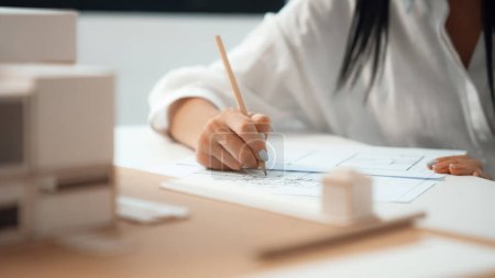 Photo for Closeup of beautiful young architect engineer hand drawing, drafting blueprint with blueprint and house model placed on working table at modern office. Focus on hand. Blurring background. Immaculate. - Royalty Free Image