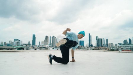 Téléchargez les photos : Danseur B-boy caucasien pratiquant la danse de rue sur le toit avec la ville ou urbaine. Prise de vue en mouvement d'un jeune homme dansant dans la rue en posant sur le gel ou en se tenant debout. Sport de plein air 2024. Endeavor. - en image libre de droit