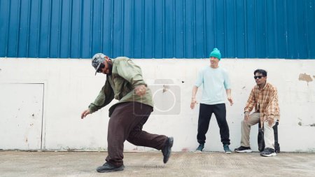 Asian hipster doing somersault in front of wall with lively friend. Attractive street dancer practicing break dance at street with blue background surrounded by friends. Outdoor sport 2024. Endeavor.