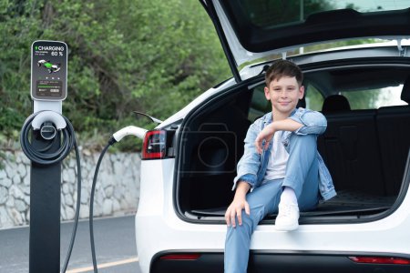 Photo for Little boy sitting on car trunk while recharging eco-friendly electric car from EV charging station. EV car road trip travel concept for alternative transportation powered sustainable energy.Perpetual - Royalty Free Image