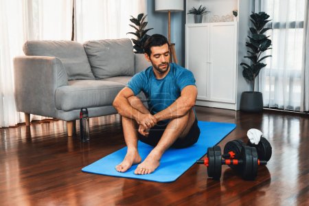 Photo for Athletic and sporty man sitting, resting on fitness mat after finishing home body workout exercise session for fit physique and healthy sport lifestyle at home. Gaiety home exercise workout training. - Royalty Free Image