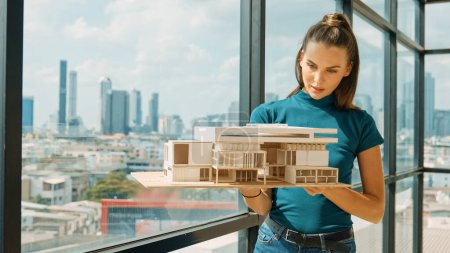 Photo for Young smart architect engineer holds architectural model while inspect house model. Professional interior designer checking house construction while standing near window with city view. Tracery - Royalty Free Image