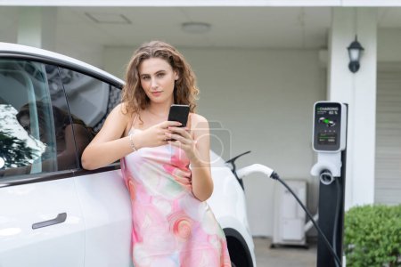 Photo for Modern eco-friendly woman recharging electric vehicle from home EV charging station. Innovative EV technology utilization for tracking energy usage to optimize battery charging at home. Synchronos - Royalty Free Image
