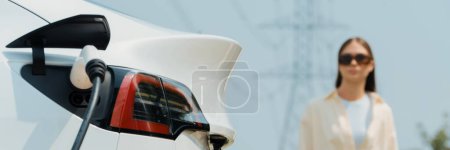 Photo for Young woman recharge EV car battery at charging station connected to power grid tower electrical industrial facility as electrical industry for eco friendly vehicle utilization. Panorama Expedient - Royalty Free Image